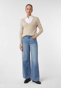 Frau steht mit einer Hand in der Tasche, trägt einen beigen V-Ausschnitt-Pullover über einem weißen Hemd, weite blaue Jeans und schwarze Schuhe vor weißem Hintergrund.