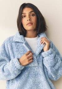 Lysebl�å fluffy fleecejakke med krave, oversized pasform og blød tekstur. Modellen justerer kraven og afslører en hvid top nedenunder.