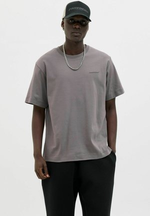 Graues kurzärmliges T-Shirt mit lässiger Passform, rundem Ausschnitt und subtilen horizontalen Details. Getragen mit schwarzen Hosen und einer Mütze.
