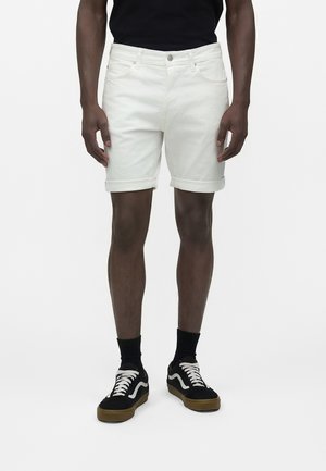 Άνδρας που φοράει λευκά σορτς με μανσέτες, μαύρες κάλτσες, ασπρόμαυρα αθλητικά παπούτσια και μαύρο πουκάμισο, στέκεται μπροστά σε απλό φόντο.