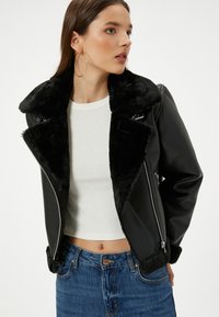 Giacca in ecopelle nera con colletto in pelliccia nera spessa, cerniere argentate e design aderente. Indossata sopra una camicia bianca e jeans blu.