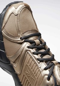 Bronzová atletická bota s černými akcenty, s texturovaným síťovaným svrškem, zpevněnou špičkou, černými šňůrkami a silnou gumovou podrážkou.