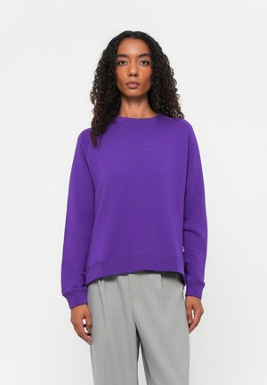Lila Sweatshirt mit Rundhalsausschnitt und langen Ärmeln, ausgestattet mit einem leicht verlängerten Saum. Graue gestreifte Hose ist unterhalb der Taille sichtbar.