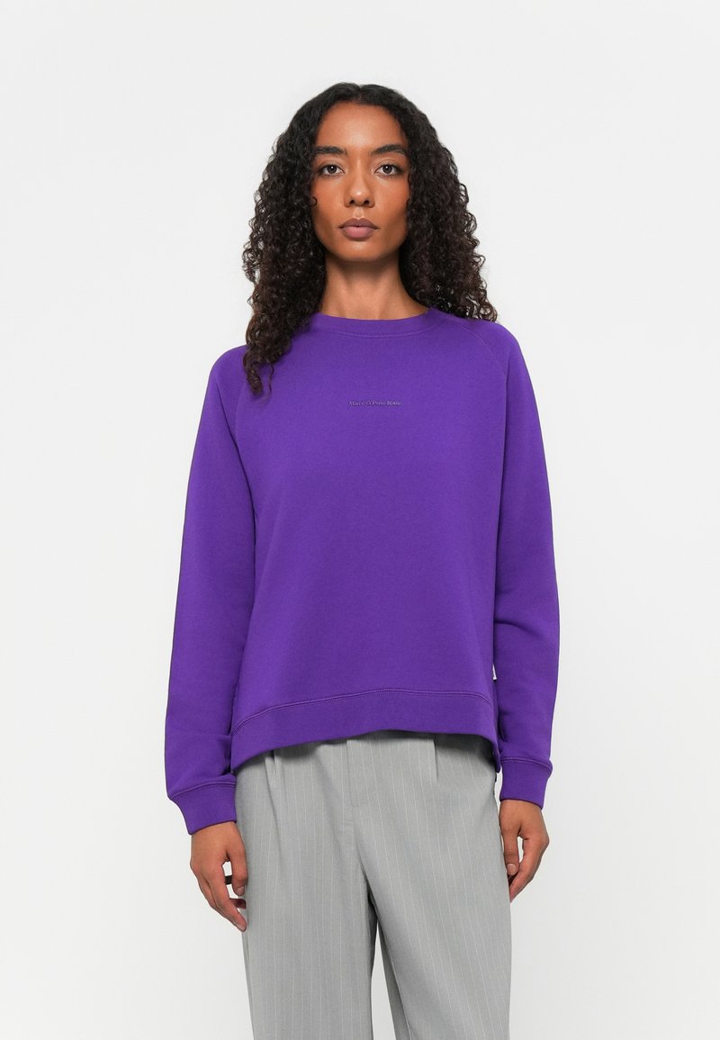 Felpa viola con collo tondo e maniche lunghe, caratterizzata da un leggero fondo smussato. Pantaloni grigi a righe visibili sotto la vita.