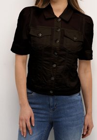 Svart kortärmad denimjacka med två bröstfickor, knäppning fram och strukturerad design. Bärs över blå jeans med slät textur.