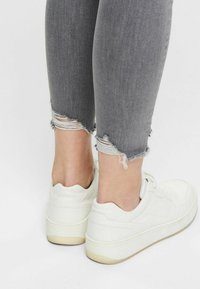 Białe sneakersy o teksturowanej powierzchni, noszone z szarymi, znoszonymi skinny jeansami z postrzępionymi końcami. Tło jest gładkie, białe.