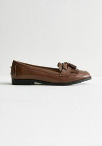 Mocasin de cuero marrón con suela plana, que presenta un borlas decorativas y detalles de costura a lo largo de la parte superior. Textura suave, diseño clásico.