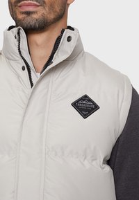 Helle beige Steppweste mit hohem Kragen, schwarzem Reißverschluss und schwarzem, rautenförmigem Logo-Patch auf der Brust. Das Material wirkt glatt.
