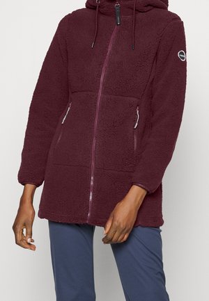Femme portant une longue veste polaire bordeaux avec capuche, fermeture éclair à l'avant, poches zippées et un écusson circulaire sur le bras gauche, associée à un pantalon bleu marine.
