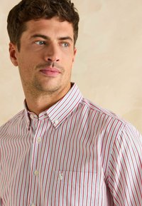 Košeľa s gombíkmi v svetlomodrej látke s vertikálnymi červenými pruhmi. Obsahuje golier, prednú vrecko a biele gombíky.