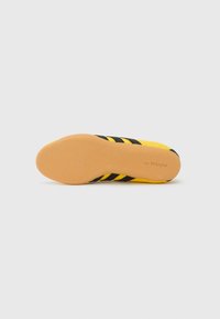 Κίτρινο αθλητικό παπούτσι με μαύρες ρίγες, λαστιχένια σόλα, υφασμάτινο επάνω μέρος με υφή και επενδεδυμένο πάτο. Λογότυπο χαραγμένο στη σόλα.