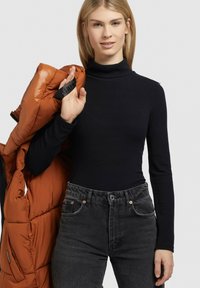 Maglione a collo alto aderente nero, abbinato a jeans grigi scuri a vita alta. Il modello tiene in mano una giacca a vento arancione lucida con una tracolla nera.