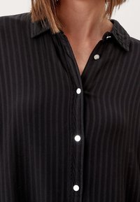 Czarna koszula zapinana na guziki, wykonana z gładkiego materiału z pionowymi paskami. Ma wskazujący kołnierzyk i srebrne napy.