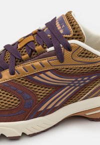 Zapatilla deportiva marrón y púrpura con tela de malla, acentos texturizados, diseños intrincados y cordones en contraste. Suela de goma con dibujo.