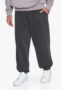 Grå sweatpants lavet af blødt stof, med en elastisk talje, ribbede manchetter og et diskret logo på låret. Sammen med hvide sneakers.