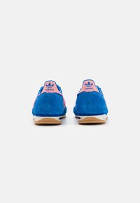 Sapatilhas de terry azul com acabamentos em rosa no calcanhar e entressola branca. Apresentam o logótipo da Adidas no calcanhar e uma superfície texturada e suave.