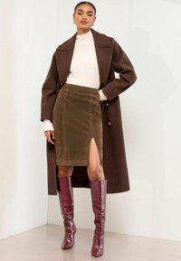 Cămașă maro cu două rânduri de nasturi, pulover crem ajustat și fustă din suede verde măsliniu cu deschidere laterală. Botine lucioase midi burgund cu model de crocodil.