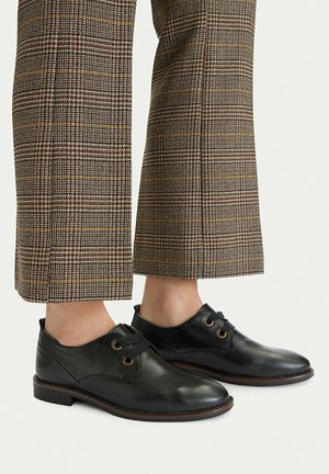 Scarpe nere in pelle con lacci indossate con pantaloni corti a quadri marroni su uno sfondo chiaro e semplice.