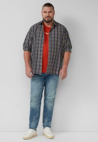 Karierte Button-Up-Bluse in Marineblau, Weiß und Braun über einem roten T-Shirt. Hellblaue Jeans und weiße Sneaker, vor einem neutralen Hintergrund stehend.