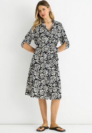 Woman wearing a black and white knee-length patterned dress with rolled-up sleeves, standing against a plain white background.