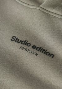 Beżowy materiał z czarnym wypukłym tekstem "Edycja studyjna" oraz współrzędnymi "55°57'03"N" wyszytymi w pobliżu szwu.