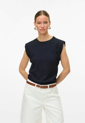 Donkerblauwe, gestructureerde mouwloze top gecombineerd met witte wijde broek en een bruine riem. Model staat met handen op de heupen tegen een effen achtergrond.