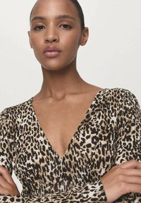 Young woman with smooth skin and pulled-back hair wears a leopard print V-neck top, standing with arms crossed against a neutral background.
