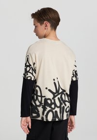 Beżowy t-shirt z krótkim rękawem z czarnym nadrukiem w stylu graffiti, nałożony na długie czarne rękawy. Luźny krój, okrągły dekolt i gładka tekstura.