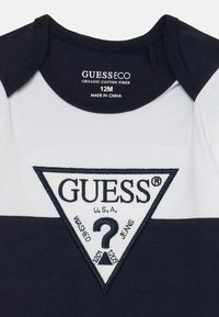 Body en coton biologique à manches courtes avec un design en blocs de couleurs blanc et bleu marine. Présente un patch logo triangulaire avec "GUESS®" et un motif de point d'interrogation.