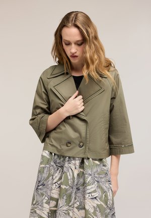 Młoda kobieta z falującymi blond włosami, ubrana w oliwkowo-zieloną krótka kurtkę i zielono-białą spódnicę w wzory, patrząca zamyślona w dół.