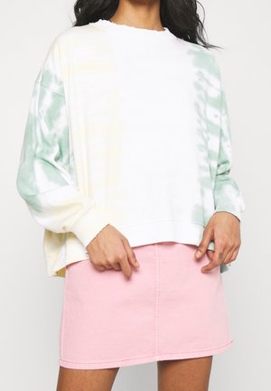 Kvinde iført en løs hvid sweatshirt med grønne og gule tie-dye ærmer og et højtkvalset pink mininederdel, stående mod en ensartet baggrund.