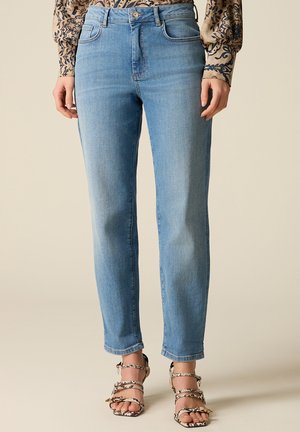 Mujer vistiendo jeans rectos de pierna ancha en azul claro, blusa de manga larga con estampado y sandalias de tacón alto con tiras y estampado de piel de serpiente.