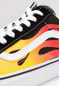 Bunter Sneaker mit gelben und schwarzen Flammenmustern, weißen Akzenten, schwarzem Wildleder-Obermaterial und weißen Schnürsenkeln vor einer strukturierten Oberfläche.