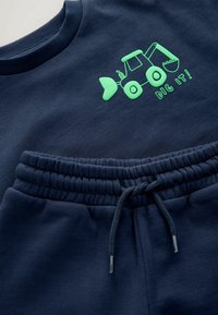Zestaw składający się z granatowej bluzy i spodni z miękkiego materiału. Na piersi znajduje się grafika zielonego traktora, a w spodniach elastyczny pas z sznurkiem.