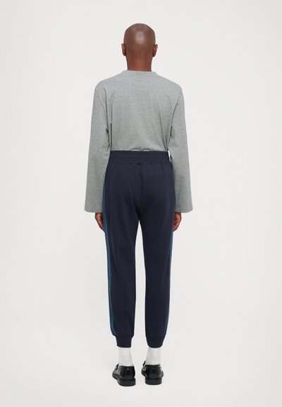 En person står med ryggen til iført en grå genser med lange ermer, marineblå joggebukser med side-stripete, hvite sokker og svarte pensko.