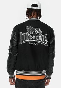Mężczyzna w czarnej kurtce varsity z skórzanymi rękawami oraz dużym białym logo "Lonsdale London" i emblematem lwa na plecach, w białej czapce noszonej tyłem.