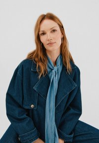 Jeansowa kurtka w ciemnoniebieskim kolorze, z dużymi klapami i jednym guzikiem. Jasnoniebieski szalik zawiązany na szyi. Modelka ma falujące brązowe włosy i kolczyki w kształcie kół.