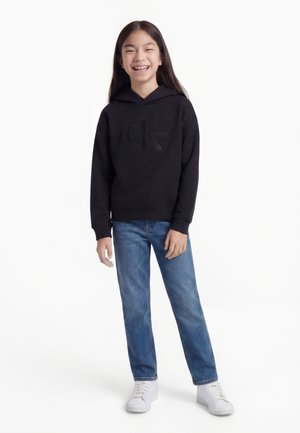Fille souriante aux longs cheveux noirs portant un sweat à capuche noir Calvin Klein, un jean bleu et des baskets blanches, debout devant un fond blanc.