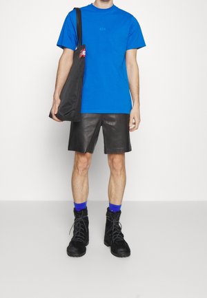 T-shirt en coton bleu avec un numéro imprimé, shorts en cuir noir, bottes noires à lacets et chaussettes bleu marine. Sac fourre-tout noir.