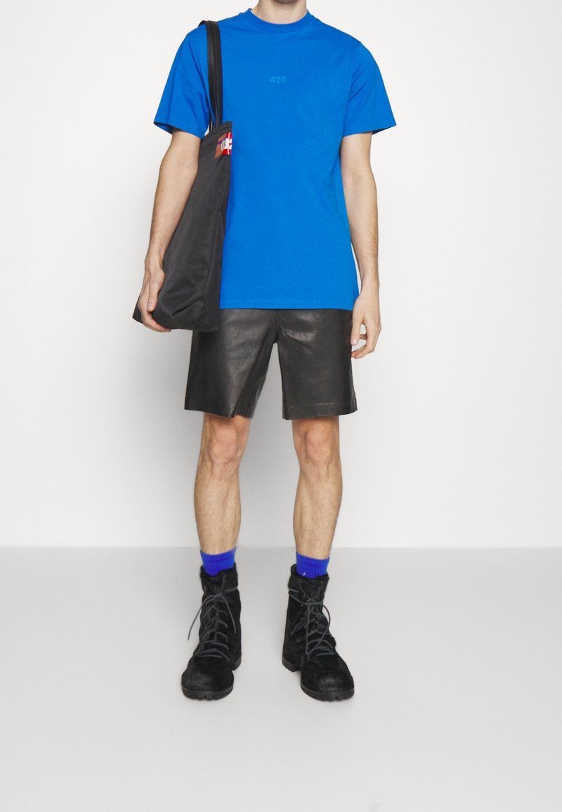 Modré bavlněné tričko s potiskem čísla, černé kožené šortky, černé šněrovací boty a modré kotníkové ponožky. Černá taška přes rameno.