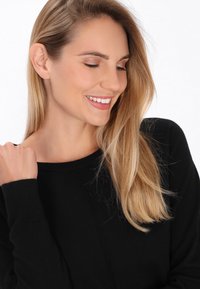 Pulover negru cu decolteu rotund, având mâneci lungi și un model texturat. Modelul are părul blond, lung și ondulat, cu un look natural de machiaj.