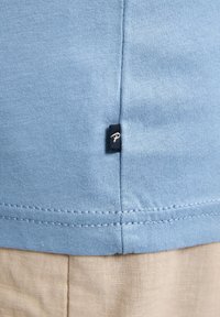 Světle modrá bavlněná látka s hladkým povrchem, vybavená malou černou etiketou s bílým "P" na švu. Jednoduchý design, kulatý výstřih.