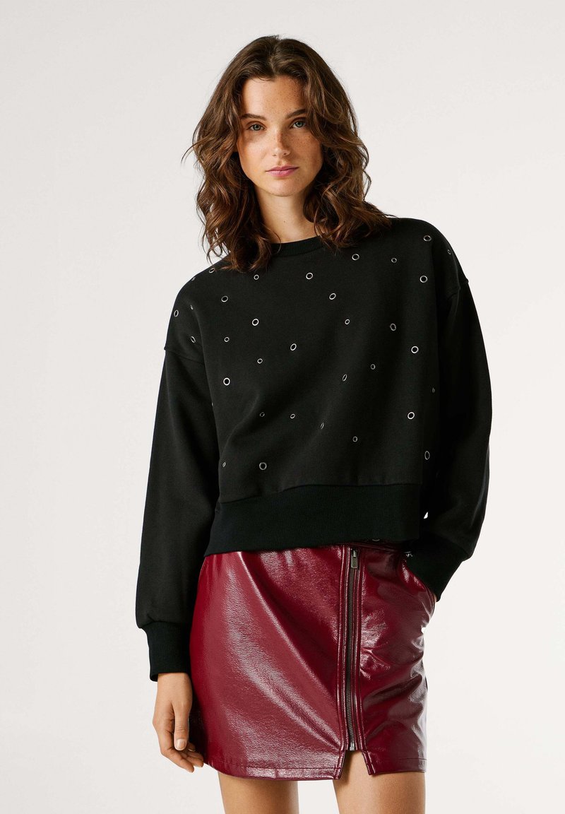 Melna džemperis ar metāla acu akcentiem, apaļu kakla izgriezumu un ribota apakšmala; kombinēts ar piegulošu, spīdīgu bordo ādas svārku ar rāvējslēdzēju.