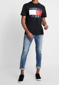 Černé bavlněné tričko s grafickým panelem v bílé, červené a modré barvě s textem "TOMMY JEANS." V kombinaci se světlými modrými tapered džíny a černými teniskami.