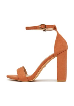 Sandalo con tacco alto in camoscio color cuoio, con cinturino alla caviglia, fibbia dorata e tacco squadrato su sfondo bianco.