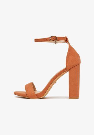 Sandalo con tacco alto in camoscio color cuoio, con cinturino alla caviglia, fibbia dorata e tacco squadrato su sfondo bianco.