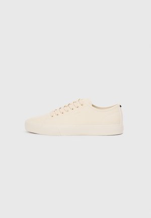 Beige canvas sneakers with round toe, flat rubber sole, and lace-up front. Features minimal design and a contrasting black accent at the heel.