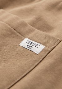 Tessuto beige con un'etichetta rettangolare bianca cucita sopra, testo recita: "Il tuo unico limite è la tua mente" e "OMBRE dal 1985".