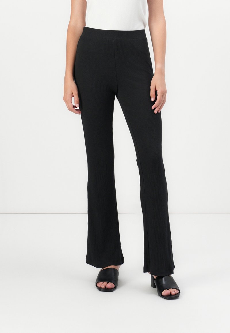 Femme portant un pantalon noir évasé, un haut sans manches blanc et des sandales à talons noires ouvertes à l'avant, debout sur fond blanc.