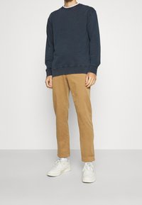 Sweatshirt bleu marine à col rond avec des poignets côtelés, associé à un pantalon chino slim beige et des baskets blanches, sur fond uni.